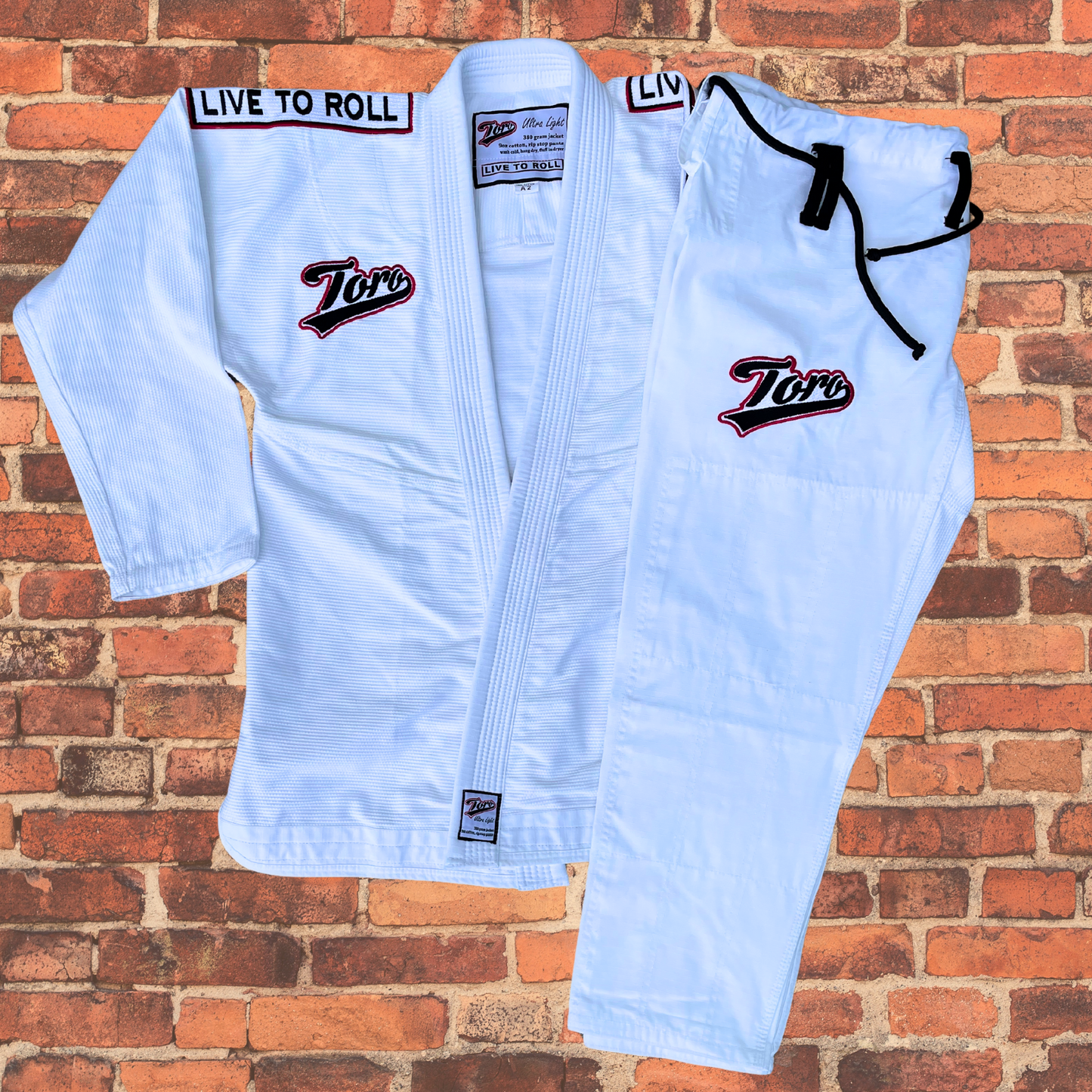 Toro's white BJJ Gi set featuring the jacket and pants with 'Live to Roll' embroidered on the shoulders and Toro logos on the chest and pants. Made from lightweight, durable material, this Gi is perfect for Brazilian Jiu-Jitsu training and competition. Displayed against a red brick wall background.