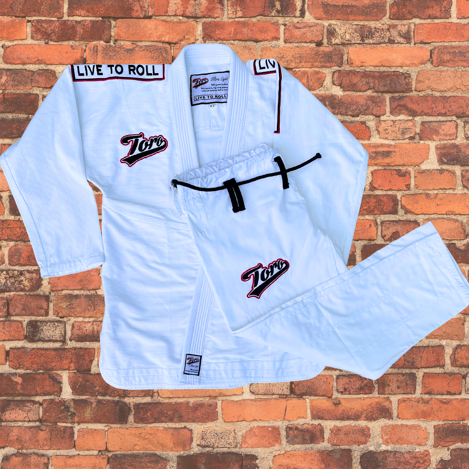 Toro's white BJJ Gi set, featuring both the jacket and pants, with 'Live to Roll' embroidery on the shoulders and Toro logos on the chest and pants. The lightweight, durable fabric makes it ideal for Brazilian Jiu-Jitsu training and competition. Displayed against a red brick wall background.