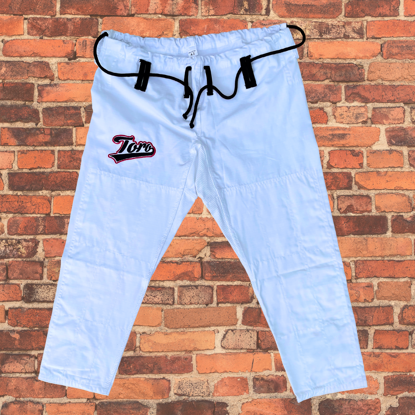 Close-up of Toro's white BJJ Gi pants featuring the Toro logo on the upper leg. The pants are equipped with a drawstring waistband for a secure fit and are made of lightweight, durable fabric, perfect for Brazilian Jiu-Jitsu training and competition. Displayed against a red brick wall backdrop.