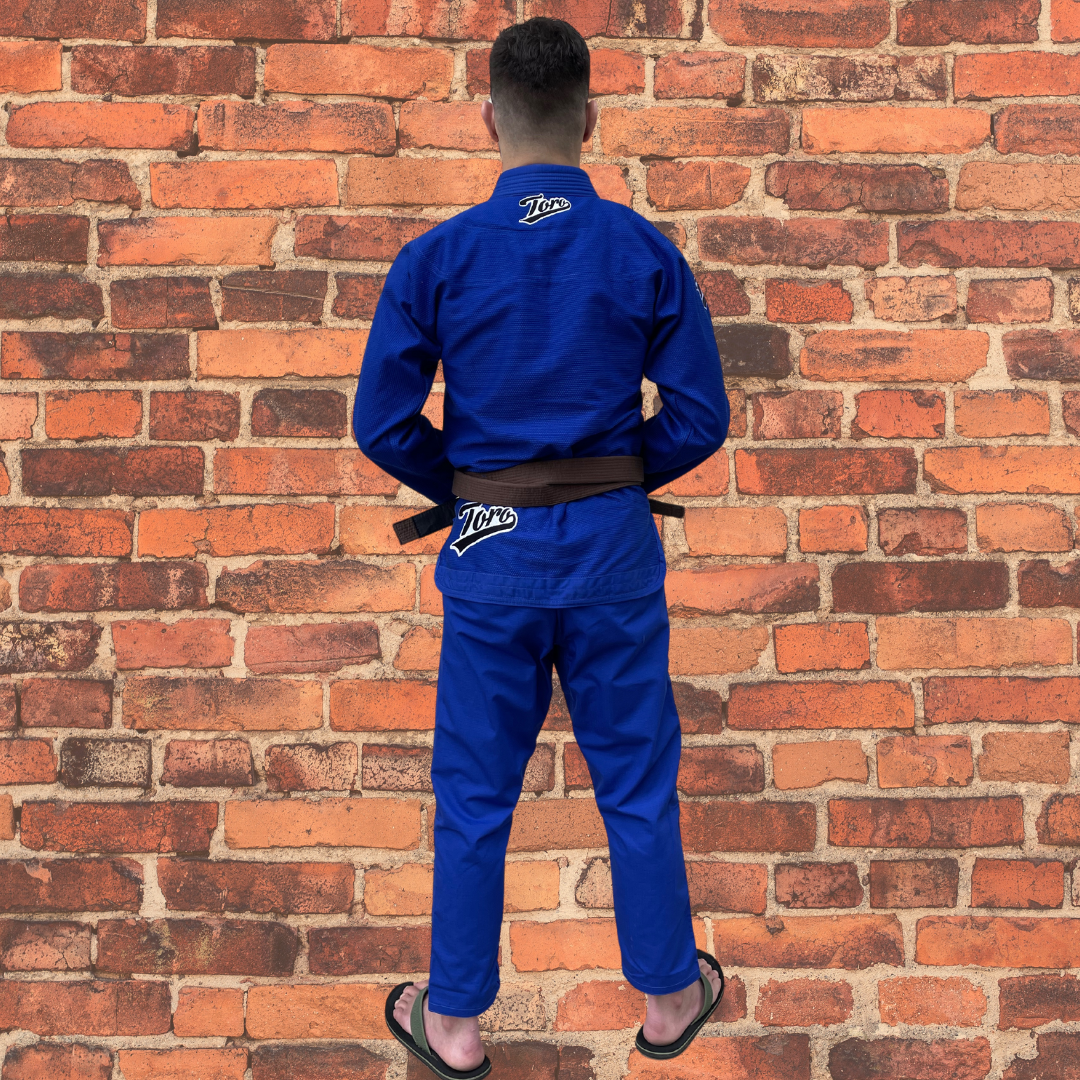 A back view of a male BJJ athlete in a blue Toro Gi standing against a brick wall, showcasing the logo on the back and pants.