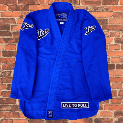 Flat lay image of the Blue Bull Toro BJJ Gi featuring embroidered Toro logos on the shoulders and chest, white "Live to Roll" patch on the lapel, blue 450-gram pearl weave material, IBJJF legal.