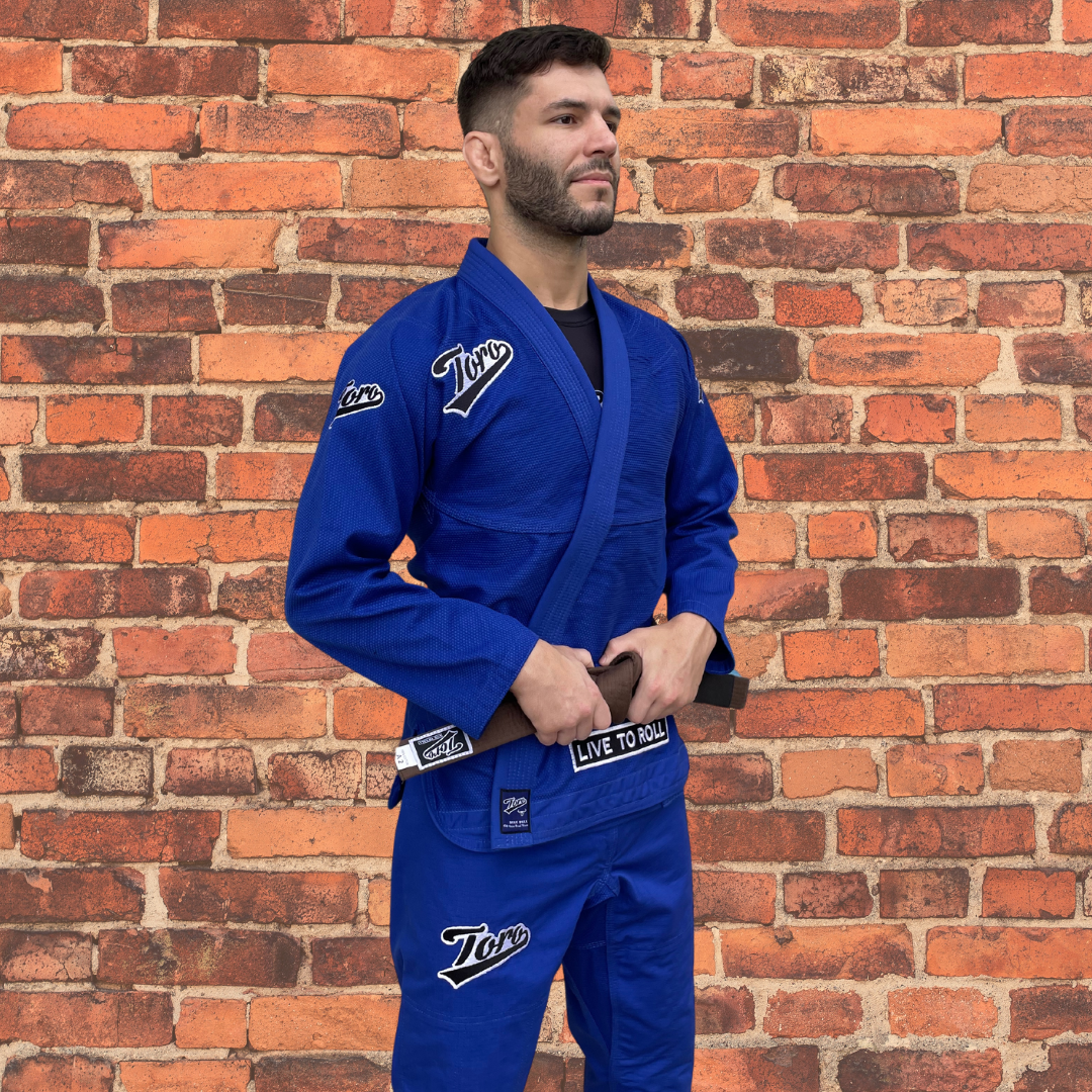 Blue Toro BJJ Blue Bull Gi with embroidered white Toro logos on the chest and shoulders, 450-gram pearl weave fabric, IBJJF legal, shown worn by a male model in front of a brick wall.