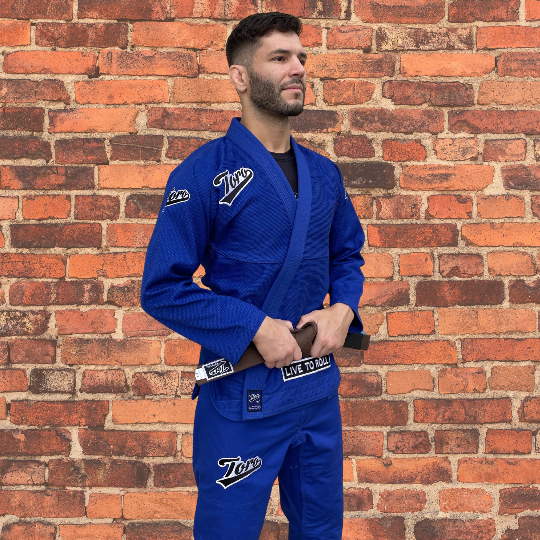 Male BJJ athlete in a blue Toro Gi with a brown belt, standing confidently against a brick wall, ready for training or competition