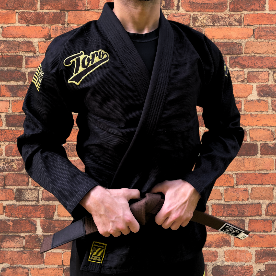 Close-up of a male BJJ athlete in a black Toro Gi with gold accents, standing confidently against a brick wall.