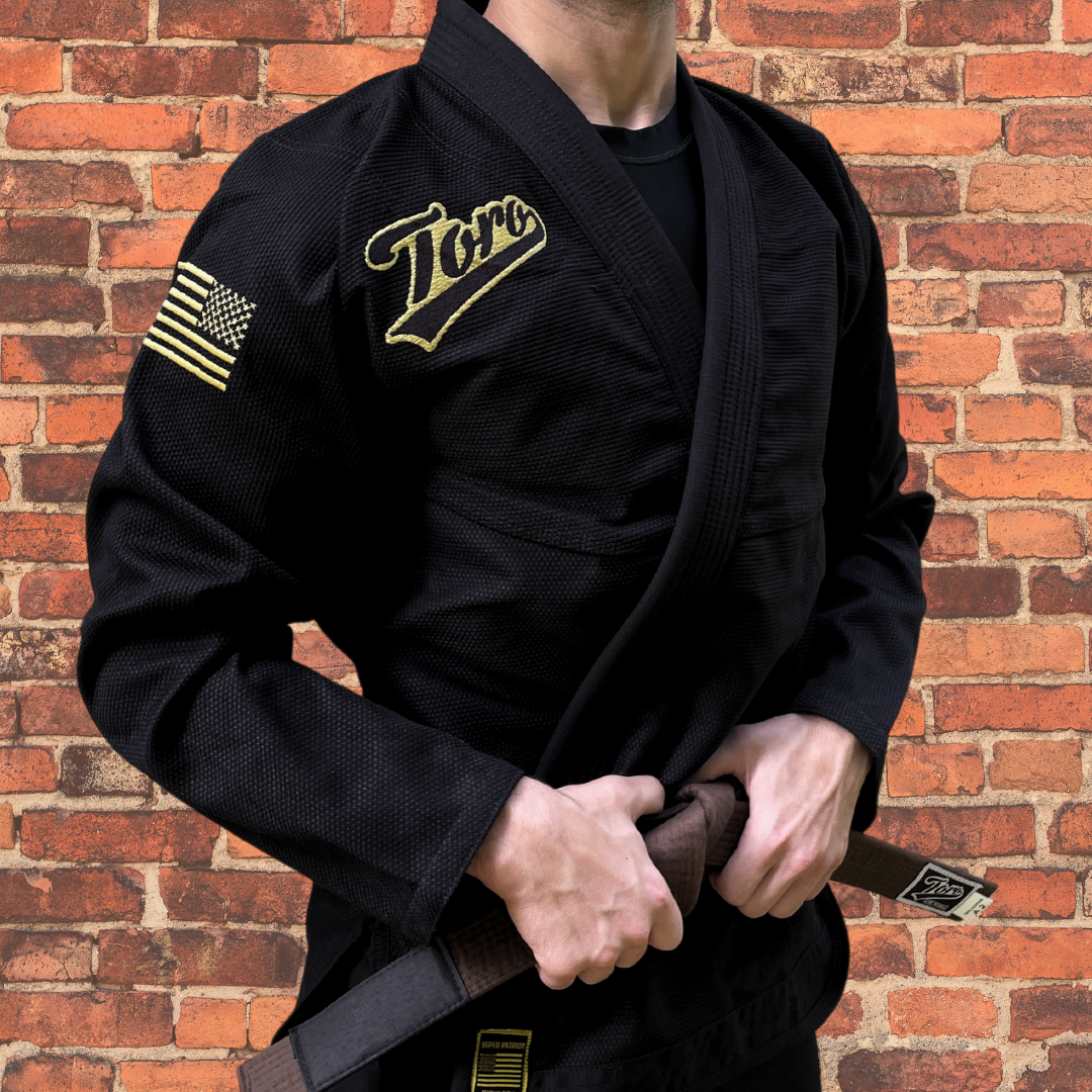 Close-up of a male BJJ athlete wearing a black Toro Gi with gold accents, standing against a brick wall, ready for competition.