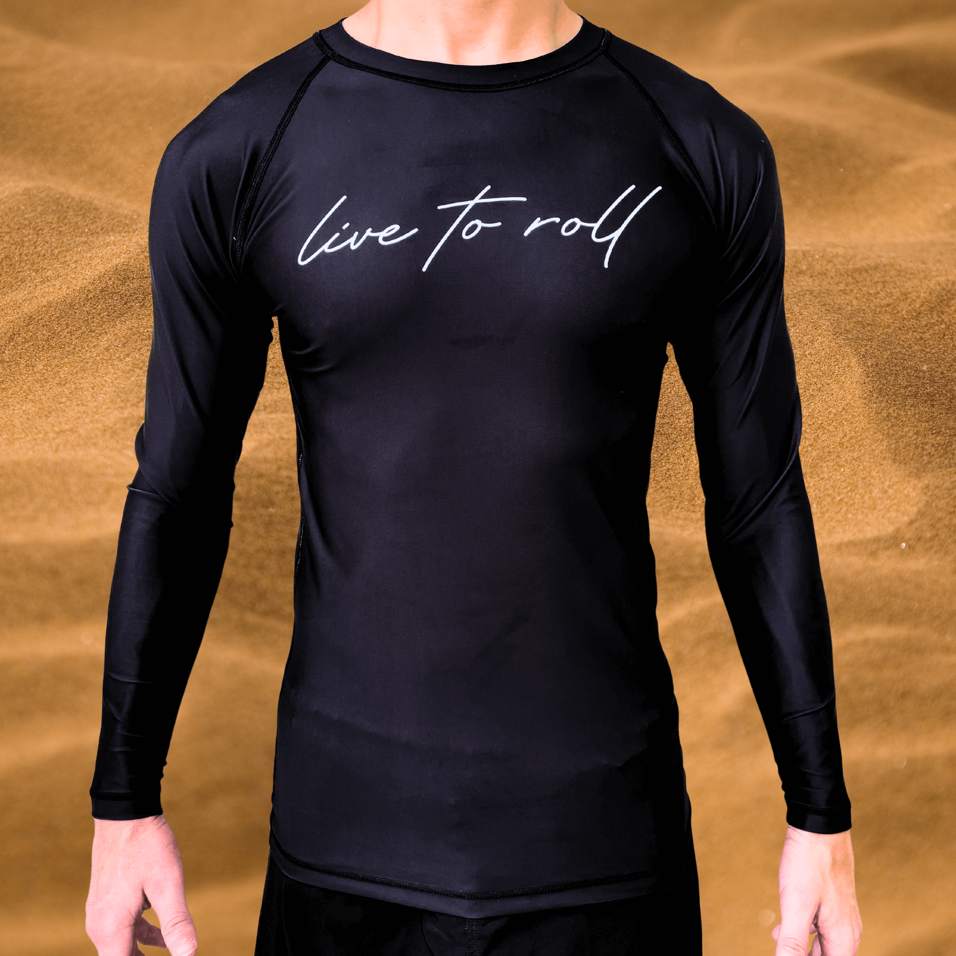 Front view of a male model wearing a black long-sleeve rash guard with “live to roll” in white script across the chest, standing in front of a sandy background.