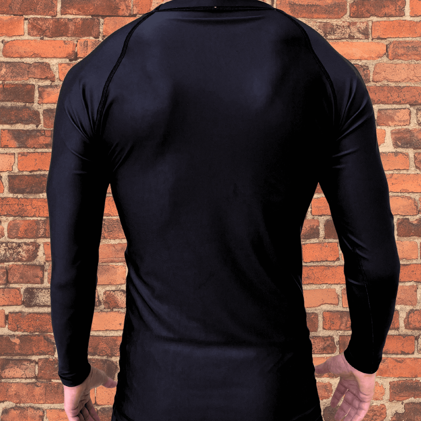 Back view of a black long-sleeve rash guard worn by a male model, with no visible design, standing in front of a brick wall.
