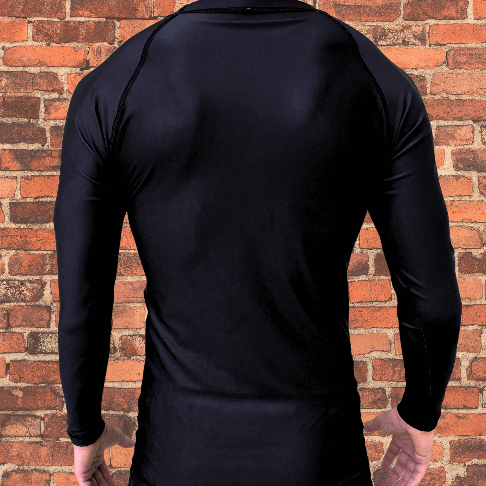 Back view of a black long-sleeve rash guard worn by a male model, with no visible design, standing in front of a brick wall.