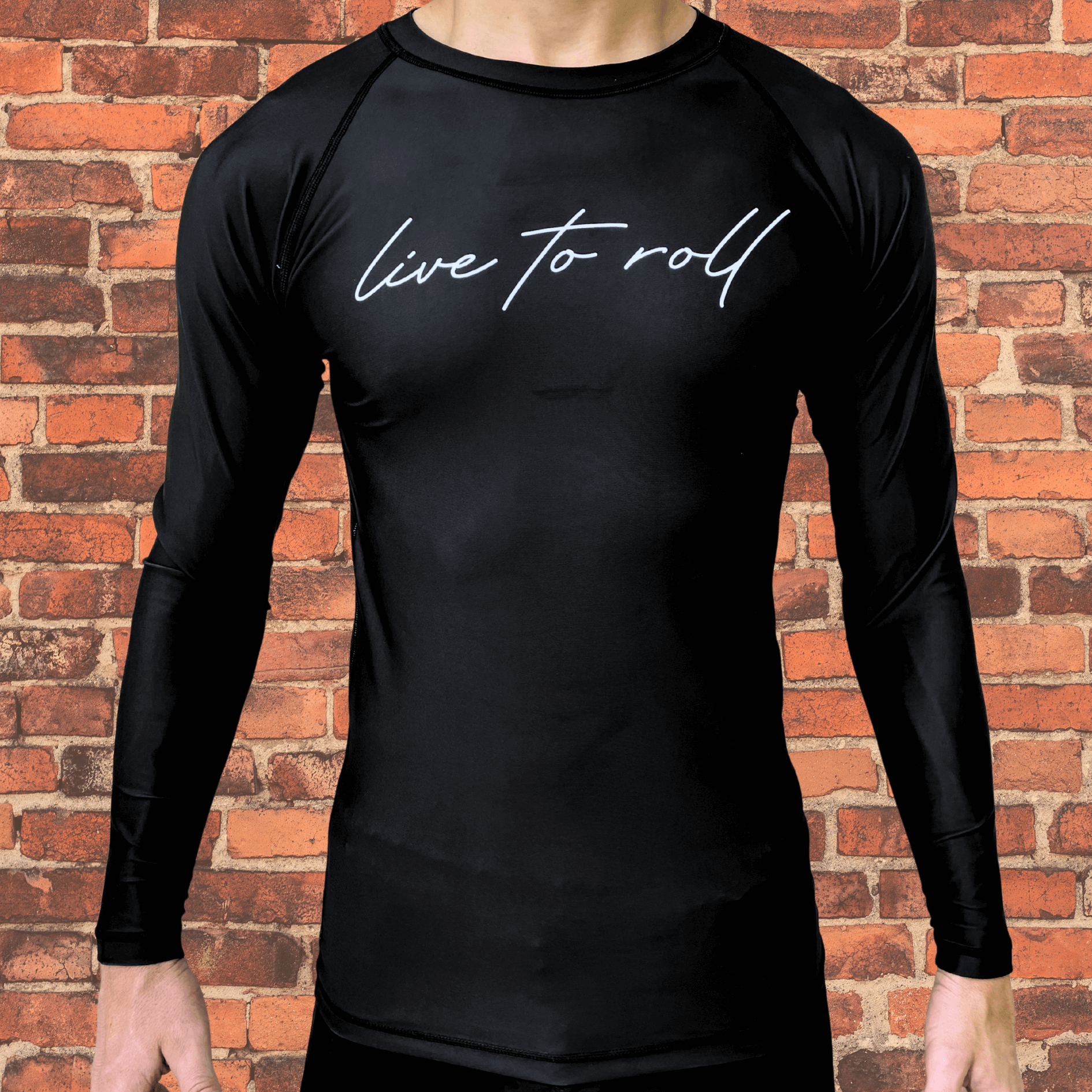 Black long-sleeve rash guard with "live to roll" script across the chest, worn by a male model standing in front of a brick wall.