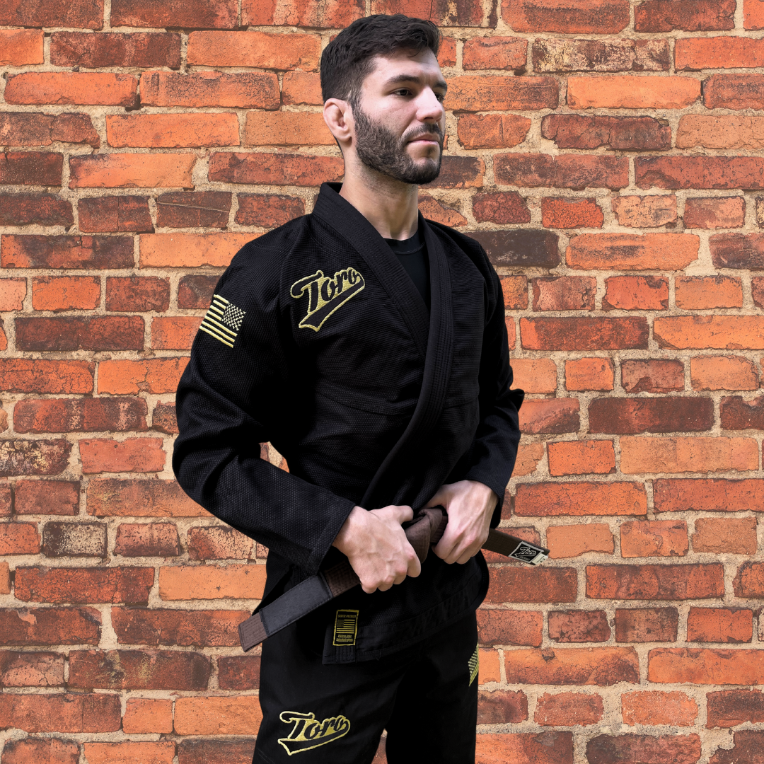 Male BJJ athlete in a black Toro Gi with gold accents and a brown belt, standing confidently against a brick wall