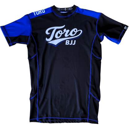 Front view of Toro 2027 blue belt rash guard with a black body, blue accents, and white chest logo