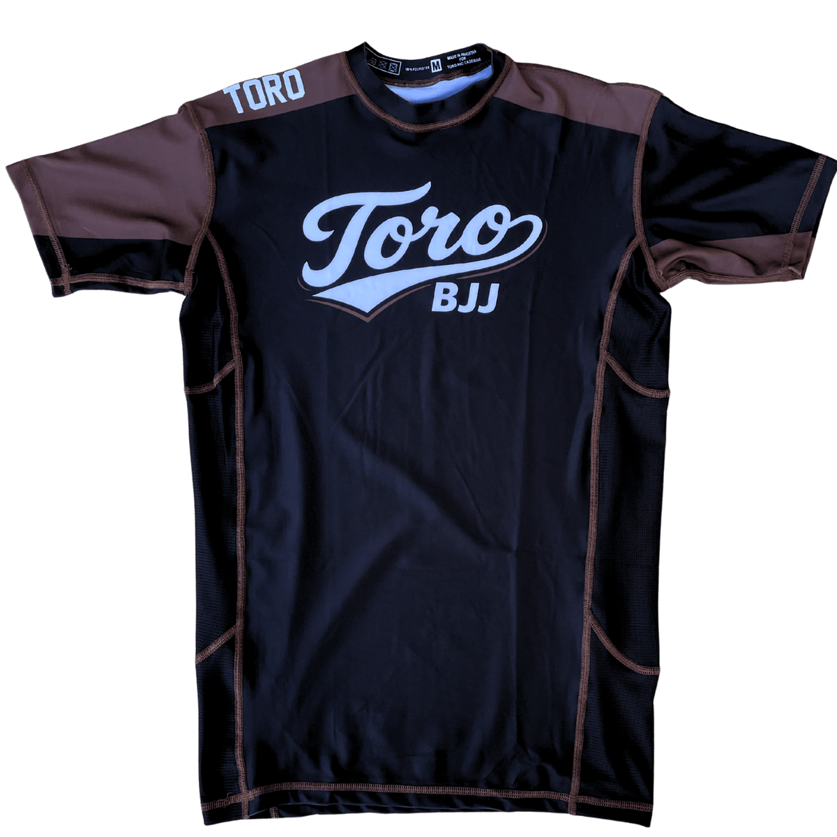 Front view of Toro 2027 brown belt rash guard with a black body, brown accents, and white chest logo