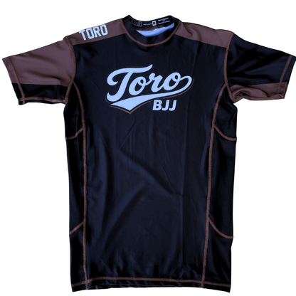 Front view of Toro 2027 brown belt rash guard with a black body, brown accents, and white chest logo