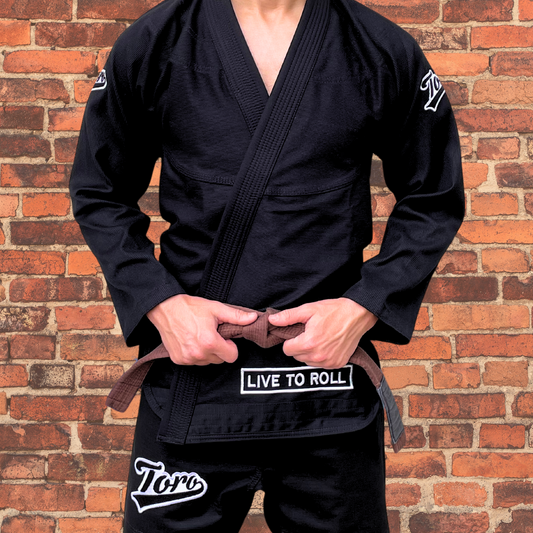 Black BJJ gi with embroidered Toro logo on shoulders, "Live to Roll" patch on gi skirt, and athlete holding brown belt, brick background.