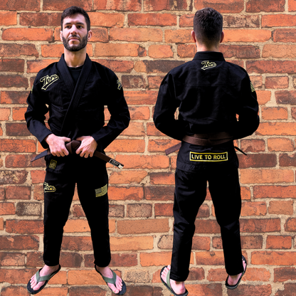 Male model wearing black BJJ gi with gold Toro logos, USA flag patch, and "Live to Roll" design on back, posing in front of brick wall.