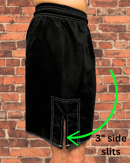 Side view of black Toro BJJ fight shorts showing 3-inch side slits with reinforced stitching, modeled against a red brick wall background.
