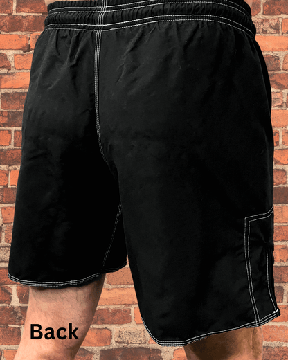 Back view of black Toro BJJ fight shorts with white contrast stitching and elastic waistband, modeled in front of a brick wall background.