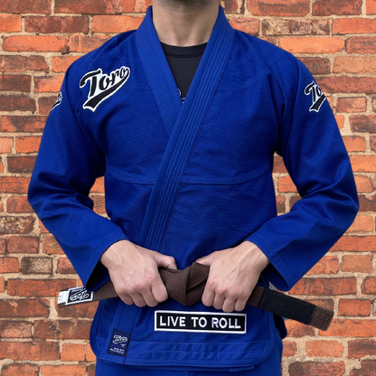 Blue Toro BJJ Gi with white embroidered logos, "Live to Roll" patch, and brown belt, displayed against a rustic brick wall.