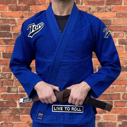 Blue Toro BJJ Gi with white embroidered logos, "Live to Roll" patch, and brown belt, displayed against a rustic brick wall.