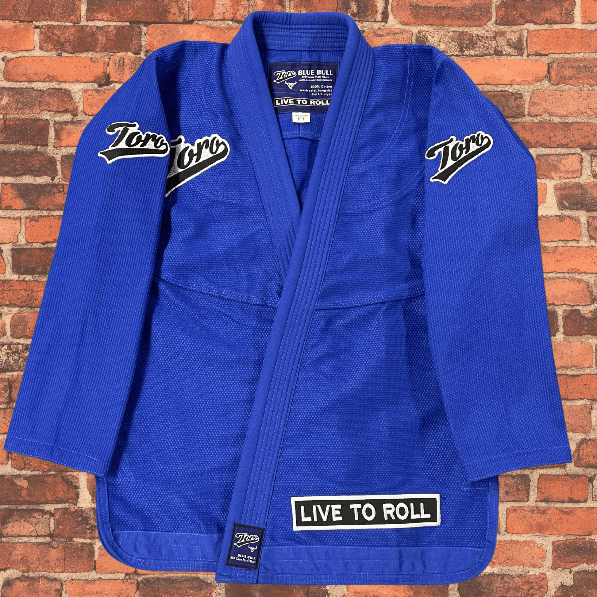 Blue BJJ Gi jacket by Toro BJJ, women’s cut, featuring "Toro" patches and "Live to Roll" text, displayed on brick background.