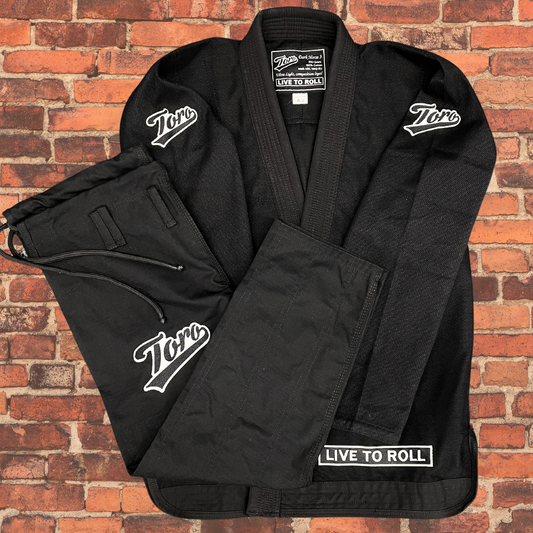 Flat lay of Toro BJJ Dark Horse 380 Black Gi with white embroidered logos and "Live to Roll" patch, displayed on brick wall background.