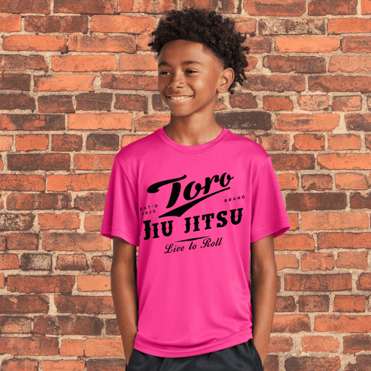 Smiling child wearing a neon pink Toro BJJ loose fit rash guard with black "Live to Roll" design, standing in front of a brick wall.