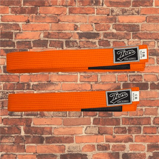 Toro BJJ kids solid orange BJJ belts in sizes M000–M5, black rank bars, Toro logo patches, displayed on brick background.
