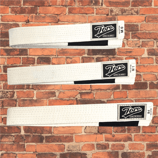 Toro BJJ kids white BJJ belts in sizes M000–M5, featuring Toro logo patch with 'Live to Roll' text, black rank bar, brick wall background.