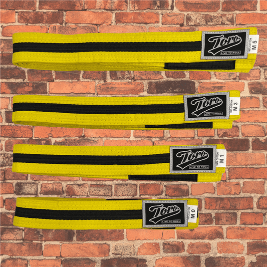 Toro BJJ kids yellow and black BJJ belts in sizes M000–M5, featuring Toro logo patches, displayed on a brick background.