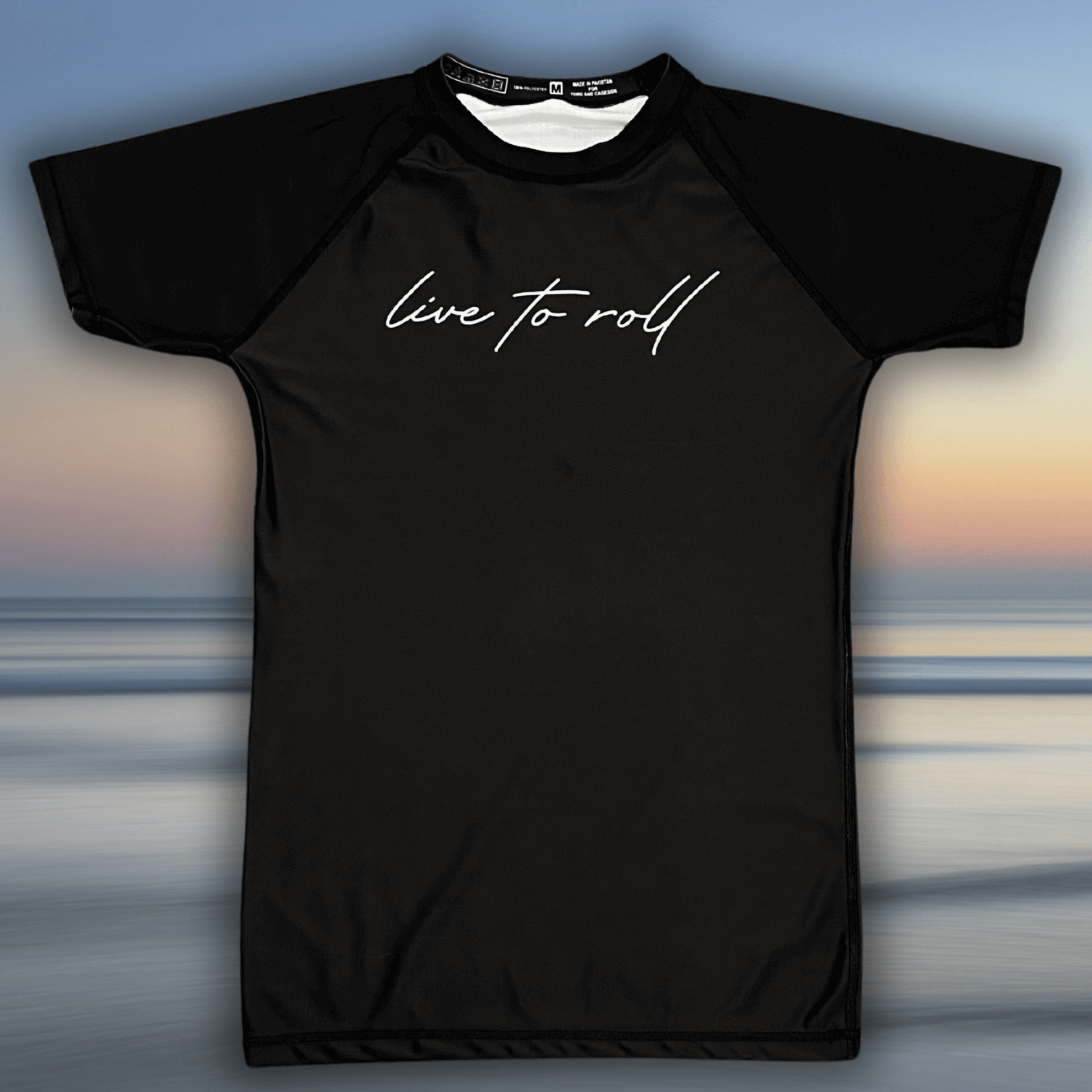 Flat lay of Toro BJJ Live To Roll black short sleeve rash guard with white script text across the chest.