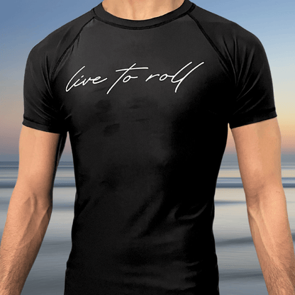 Man wearing the Toro BJJ Live To Roll black rash guard with white script text, standing in front of a beach sunset background.