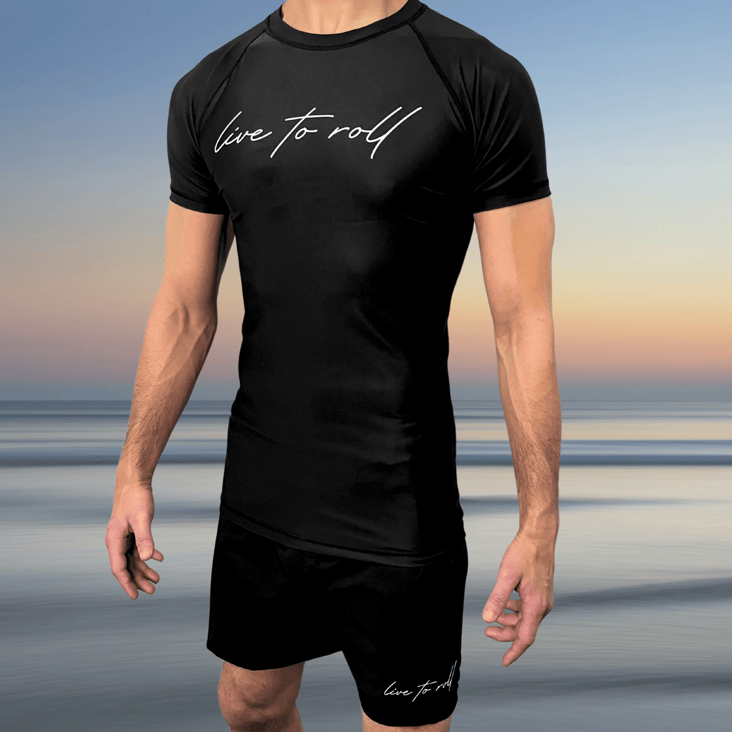Male athlete wearing Toro BJJ’s Live To Roll black rash guard and shorts set with white script logo, standing in front of a beach sunset.