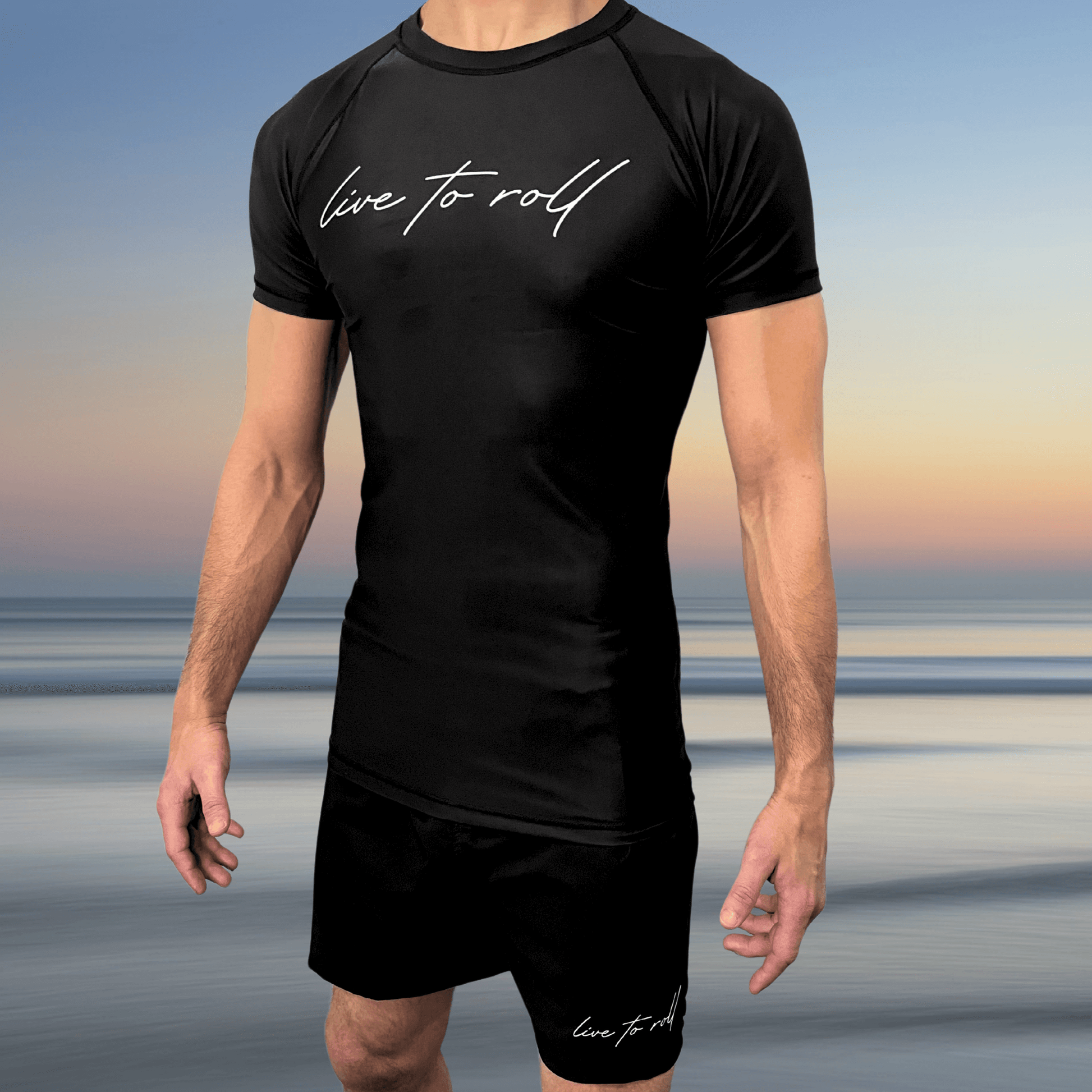 Male athlete wearing Toro BJJ’s Live To Roll black rash guard and shorts set with white script logo, standing in front of a beach sunset.