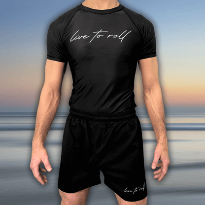 Male model wearing Toro BJJ’s Live To Roll NoGi rash guard and shorts set with white handwritten-style logo on black, standing against a beach background.
