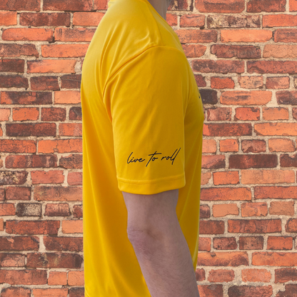 Close-up of Toro BJJ Loose Fit Rash Guard in gold, sleeve detail with "Live to Roll" text, side view against brick wall background.