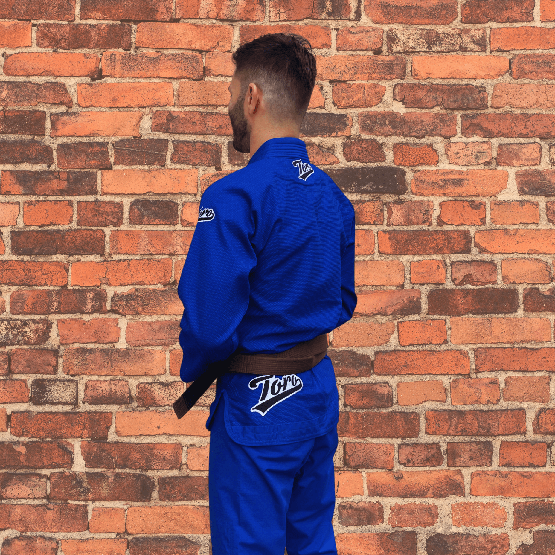Male athlete wearing Toro BJJ Men’s Blue Bull Gi with brown belt, Toro patches on shoulders and back flap, brick wall background.
