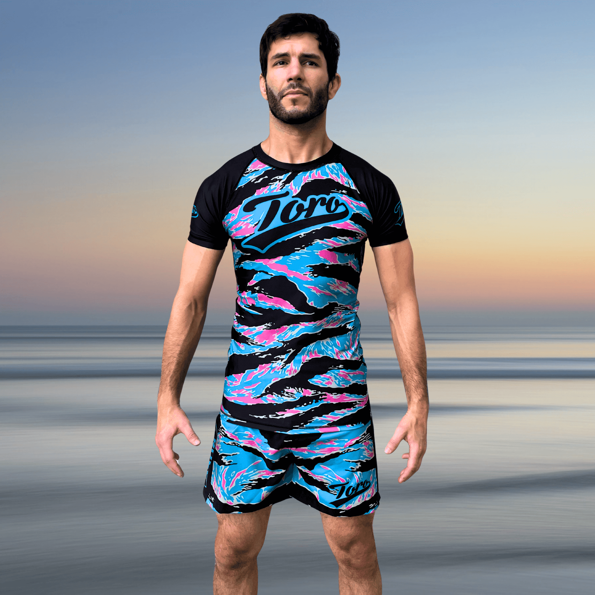 Athlete wearing Toro BJJ Miami Camo NoGi kit featuring bold blue, pink, and black camo pattern with matching rash guard and shorts, standing in front of a beach sunset background
