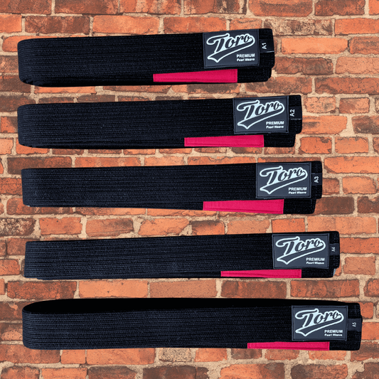 Premium black BJJ belt with a red bar, Toro logo patch, and durable pearl weave, displayed on a rustic brick wall background