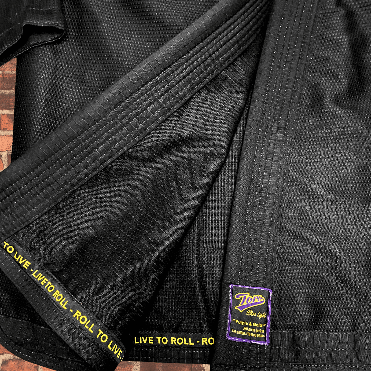 Closeup of black BJJ Gi fabric with Toro purple and gold patch and Live to Roll trim, IBJJF approved Purple & Gold Gi