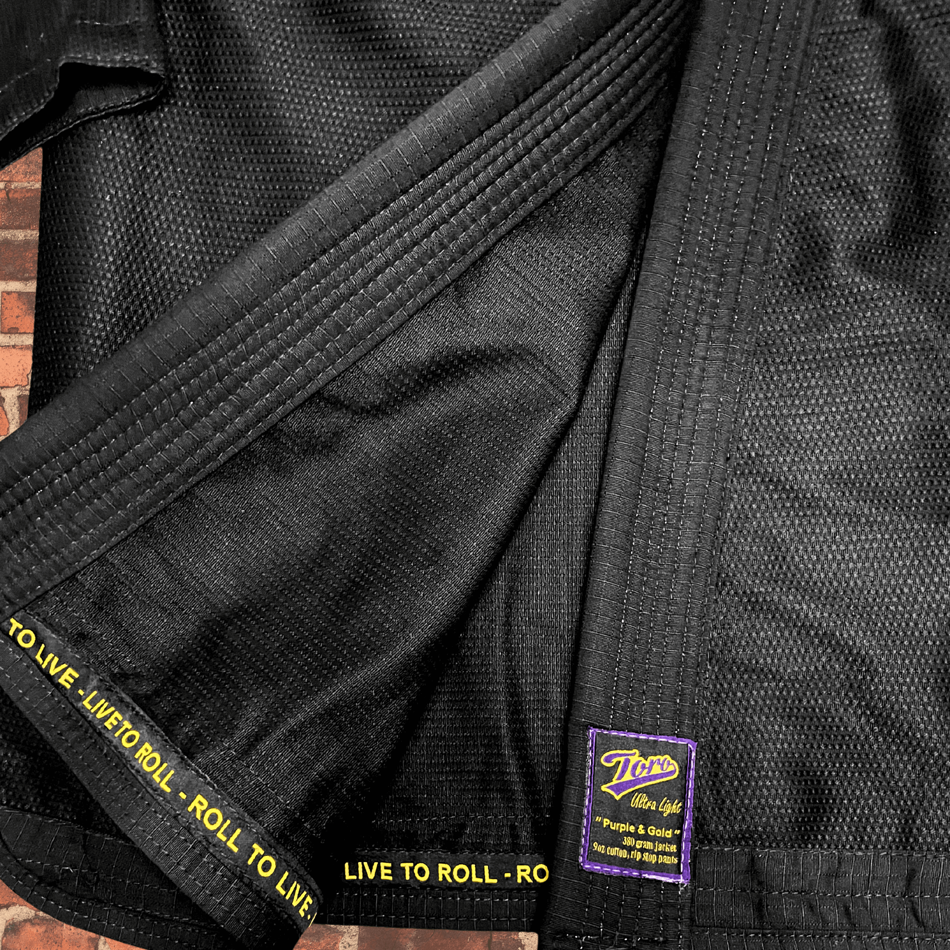 Closeup of black BJJ Gi fabric with Toro purple and gold patch and Live to Roll trim, IBJJF approved Purple & Gold Gi