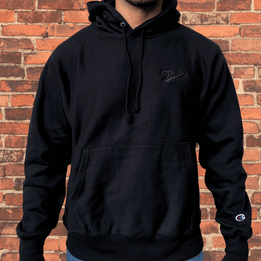 Model wearing Toro x Champion Reverse Weave Hoodie black on black with embroidered Toro logo and Champion logo on sleeve.