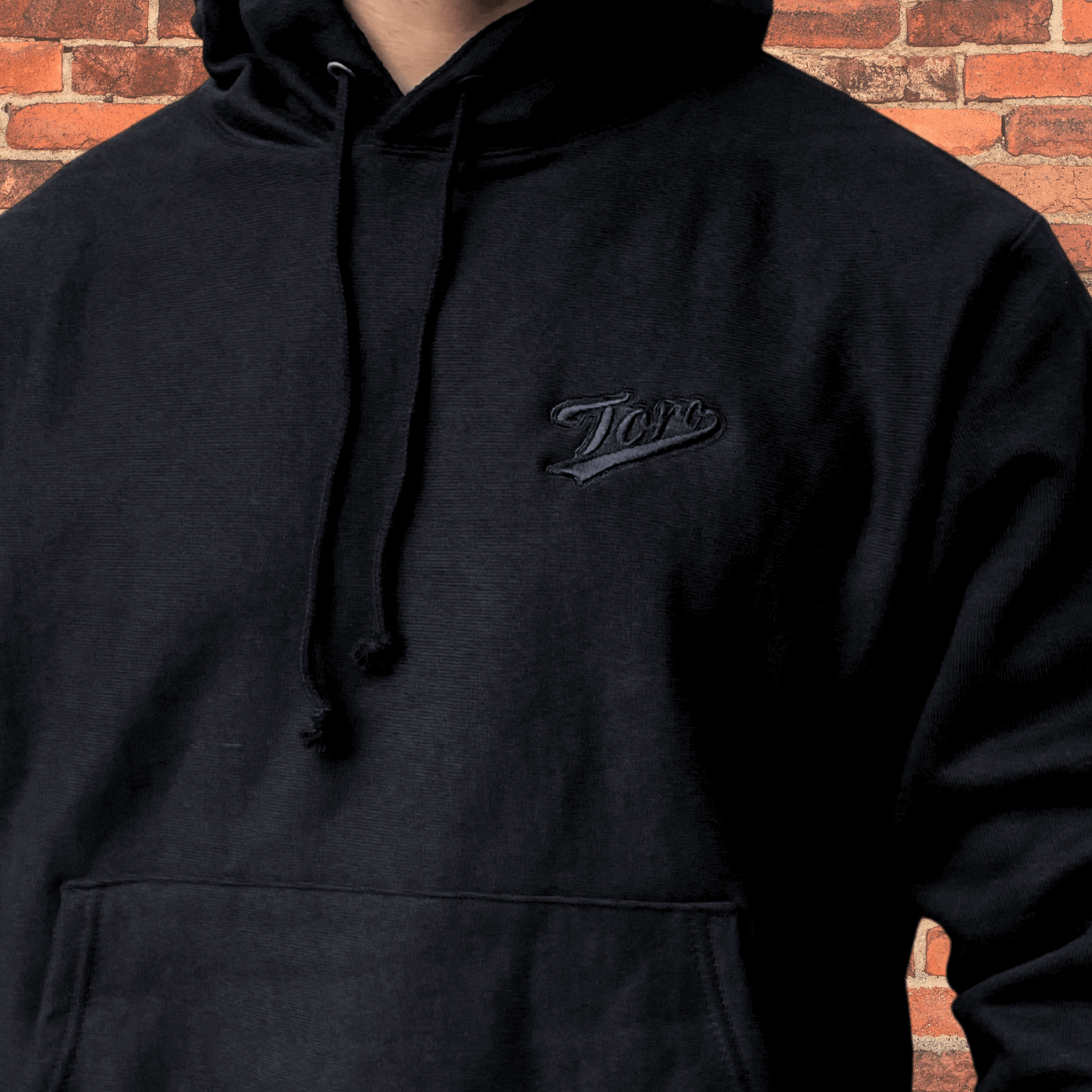 Close-up of embroidered Toro logo on black Champion Reverse Weave Hoodie showing tonal black stitching.