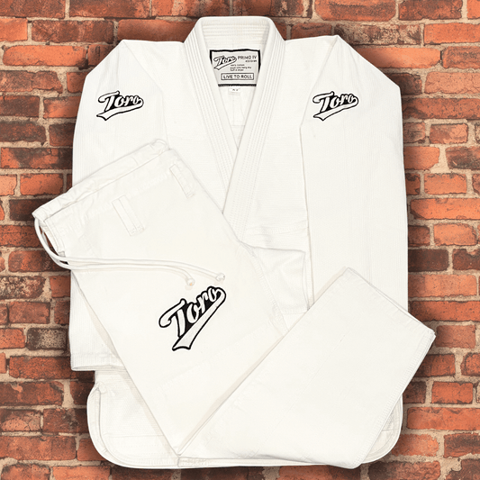 Back view of the Toro Primo White BJJ Gi jacket displayed flat against a brick wall background, featuring "Live to Roll" patch and shoulder logos with text highlighting 450 gram comp legal gi.