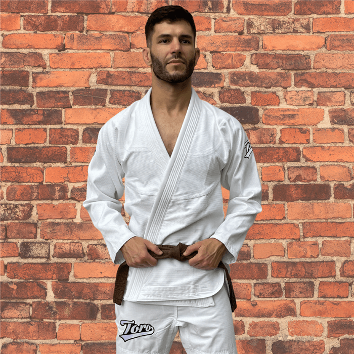 Male BJJ athlete wearing the Toro Primo White BJJ Gi with a brown belt, standing against a brick wall.