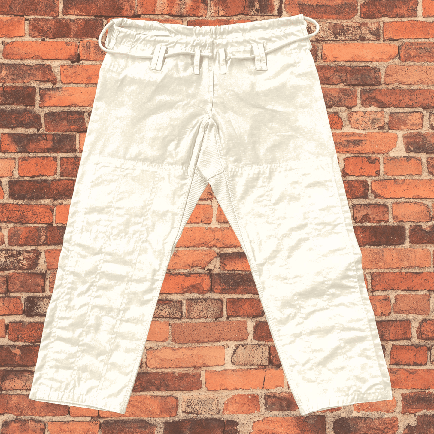 Front view of white Toro BJJ gi pants featuring reinforced knees, belt loops, and a drawstring closure, displayed against a red brick wall.