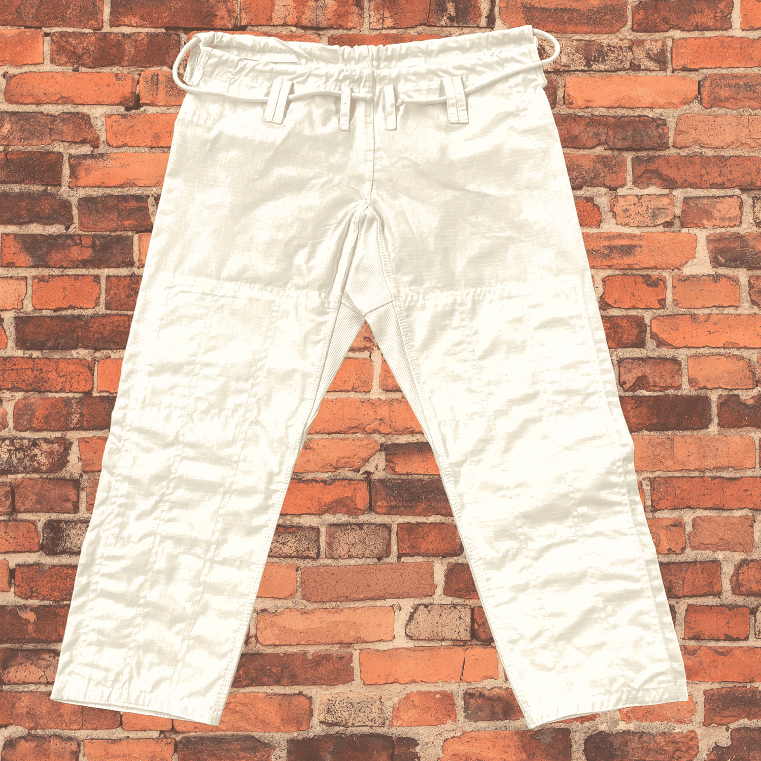 Front view of white Toro BJJ gi pants featuring reinforced knees, belt loops, and a drawstring closure, displayed against a red brick wall.